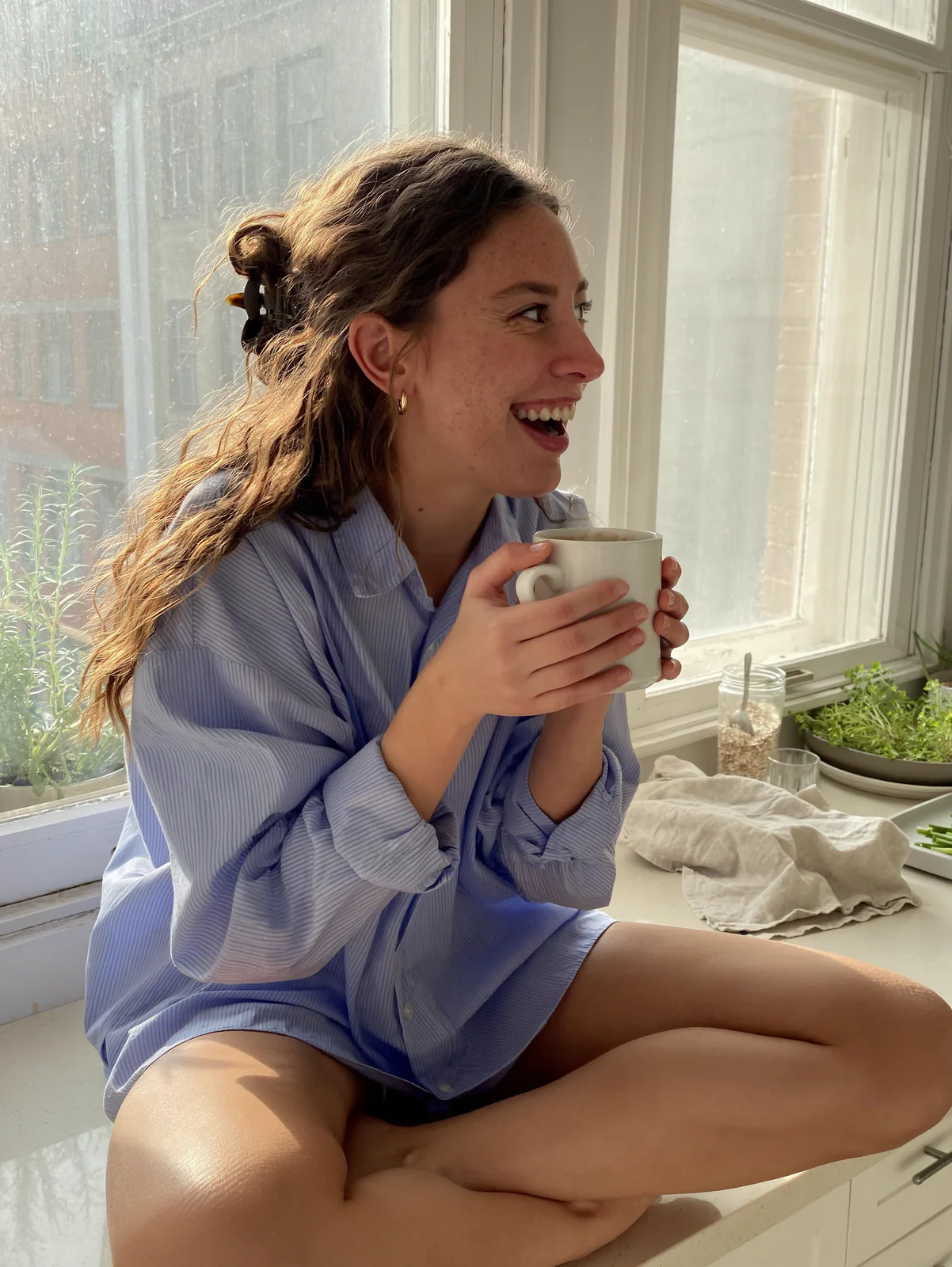MOSS Image candid iPhone photo of a woman laughing with a coffee mug on a sunny kitchen windowsill